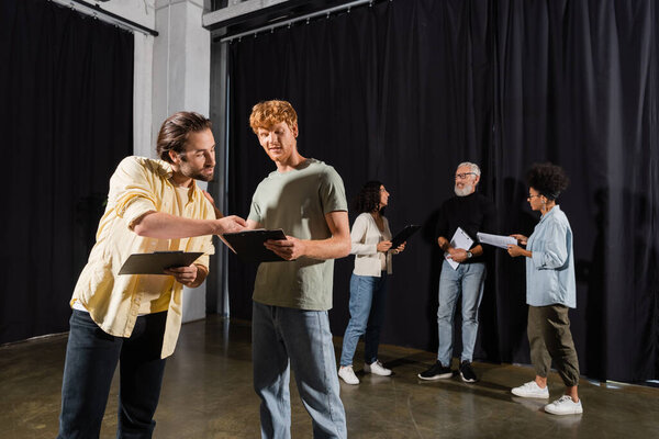 young actors holding clipboards with screenplays near interracial theater troupe on background