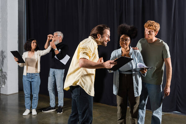 smiling man showing screenplay to interracial actors near art director and multiracial woman rehearsing on background