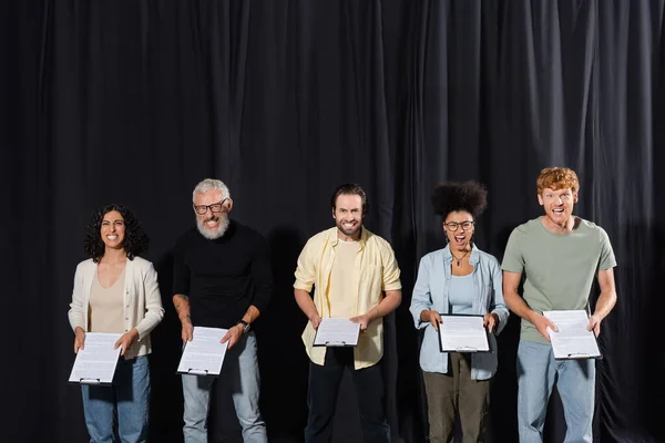 multicultural actors with bearded screenwriter holding clipboards and grimacing at camera during acting skills lesson