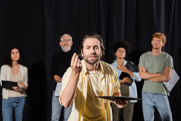 emotional man with clipboard looking at camera and gesturing during rehearsal near multiethnic actors and producer on blurred background
