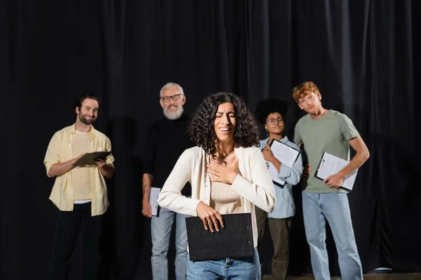 smiling multiracial woman holding clipboard and smiling near interracial actors and art director on blurred background
