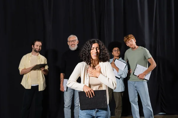 stock image smiling multiracial woman holding clipboard and smiling near interracial actors and art director on blurred background