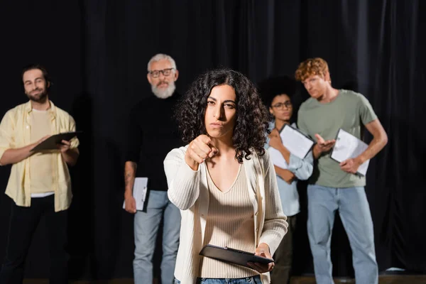 multiracial woman with serious face expression pointing at camera while rehearsing near blurred actors and mature art director