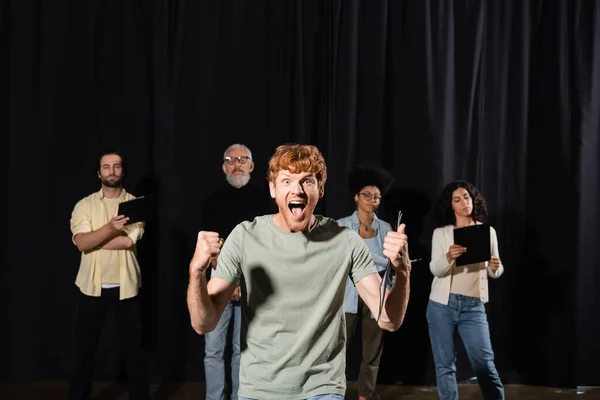 emotional redhead man showing thumb up and screaming near multiethnic students and teacher of acting skills on background
