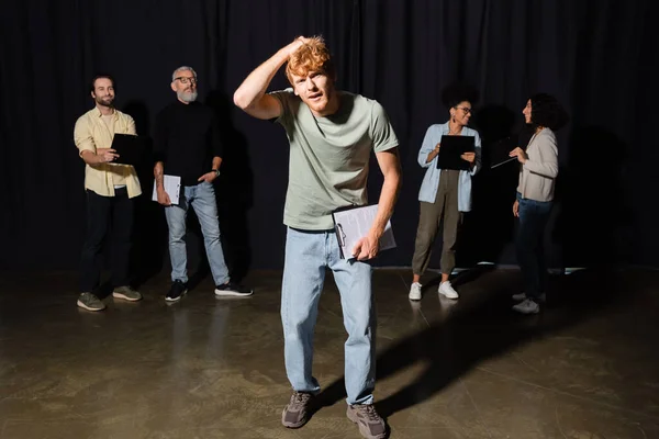 confused redhead man holding clipboard and touching head near multiethnic students and teacher of acting skills