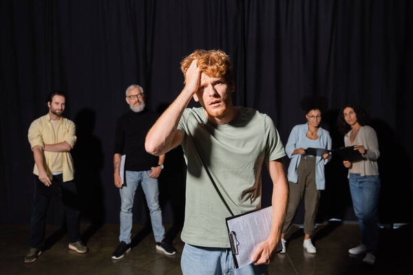 frustrated redhead man holding clipboard and touching head while looking at camera near actors and producer on blurred background