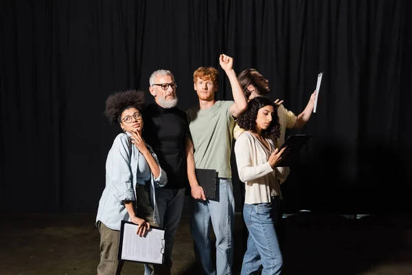 stock image redhead man looking at camera while standing with raised hand near multiethnic actors and bearded producer in theater