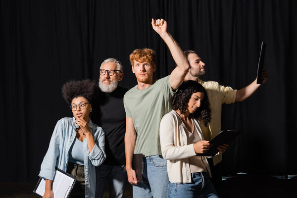 redhead man standing with raised hand near bearded art director and interracial actors with clipboards
