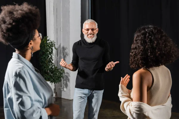 cheerful grey haired man pointing with thumb near multiethnic actresses in acting skills school