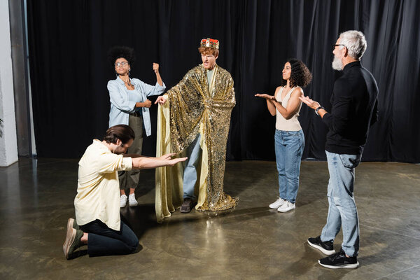 redhead man in king costume rehearsing role near multiethnic actors and art director in acting school