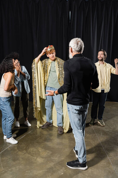 redhead man adjusting king crown near cheerful interracial actors and grey haired producer gesturing on foreground