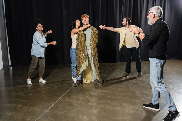 grey haired art director gesturing near interracial students rehearsing scene of king assassination in theater