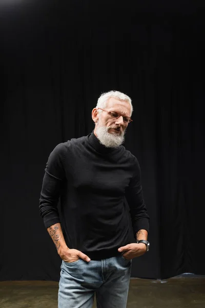 stock image mature tattooed actor in eyeglasses and black turtleneck standing with hands in pockets of jeans in theater