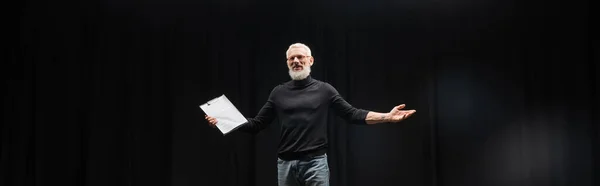 Stock image grey haired tattooed man in black turtleneck holding screenplay while rehearsing in theater, banner. Translation of tattoo: kanji, danger