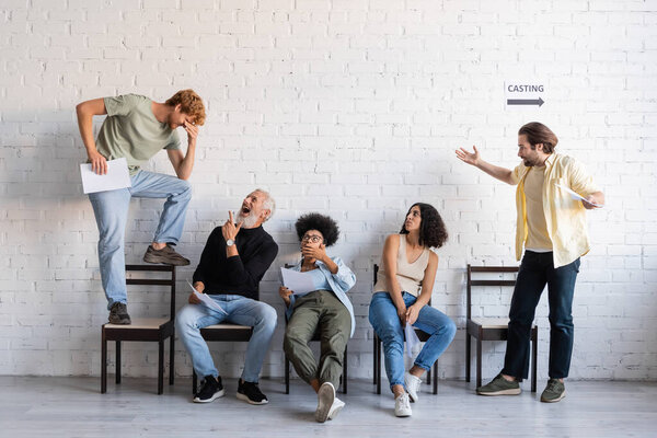 bearded man laughing and pointing at redhead man standing on chair near interracial actors waiting for casting