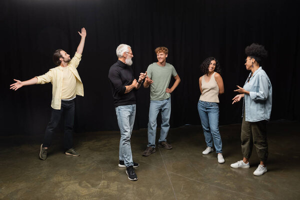young woman posing with outstretched hands near grey haired art director talking to interracial actors. Translation of tattoo: om, shanti, peace