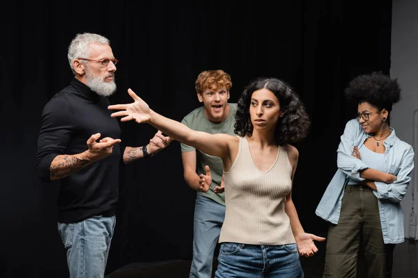 stock image multiracial woman standing with outstretched hand near bearded art director and emotional interracial students in acting school