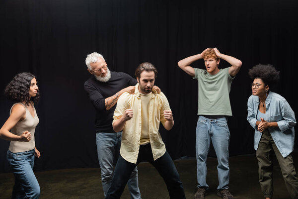 bearded acting skills teacher touching shoulders of young man during rehearsal near multiethnic actors. Translation of tattoo: om, shanti, peace