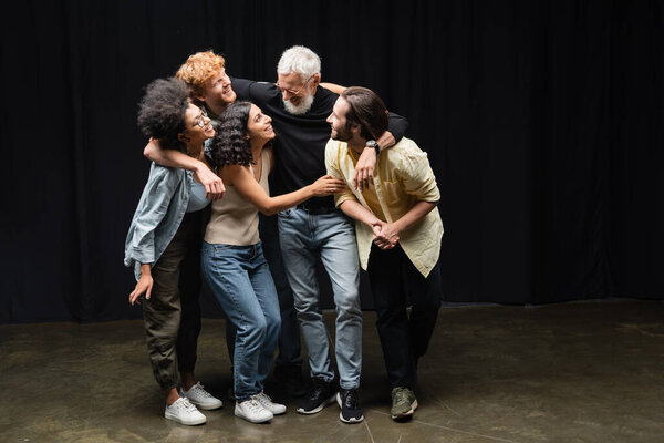 full length of young multicultural actors embracing bearded producer on stage in theater