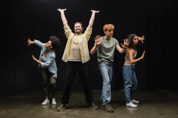 stock image young man standing with raised hands and closed eyes near multiethnic students rehearsing on stage in theater