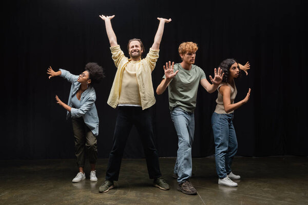 young man standing with raised hands and closed eyes near multiethnic students rehearsing on stage in theater