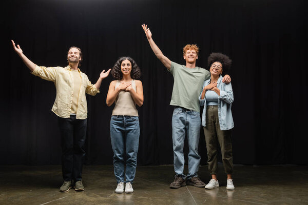 full length of happy multiethnic actors standing with raised hands on scene of theater