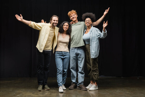 full length of cheerful interracial actors embracing while waving hands and smiling at camera in theater