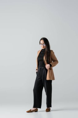 full length of asian businesswoman in beige blazer and black pants standing with hand in pocket on grey  clipart