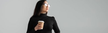 brunette asian woman in stylish black turtleneck holding paper cup while looking away isolated on grey, banner 