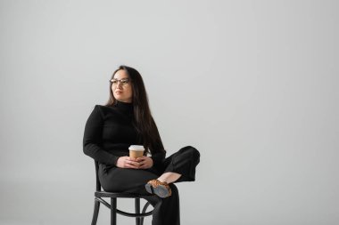 brunette asian woman in black outfit and glasses holding paper cup while sitting on chair isolated on grey 