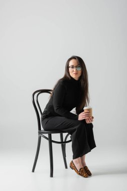 full length of brunette asian woman in black outfit and glasses holding paper cup while sitting on chair on grey 