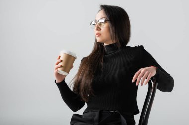 asian woman in black turtleneck and glasses holding paper cup while sitting on chair isolated on grey 