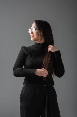 smiling asian woman in black turtleneck and glasses looking away isolated on dark grey  clipart