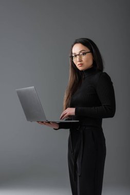 pretty asian freelancer in black turtleneck and glasses holding laptop on dark grey 