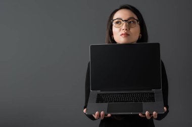 pretty asian woman in black turtleneck and glasses holding laptop with blank screen isolated on dark grey 