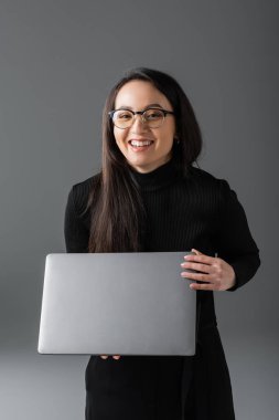 joyful asian woman in black turtleneck and glasses holding laptop on dark grey 