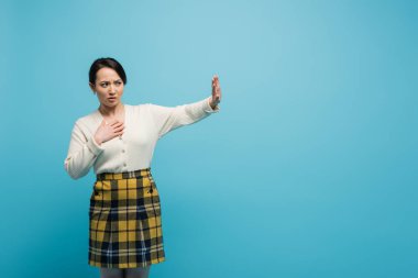 young asian woman in plaid skirt standing with outstretched hand while showing stop isolated on blue 