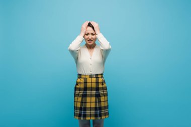 worried young asian woman in checkered skirt holding hands on head isolated on blue  clipart