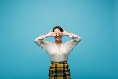 Young brunette woman smiling and covering eyes isolated on blue 
