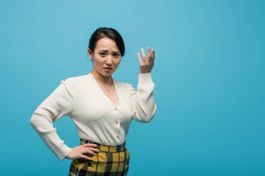 Confused asian woman in cardigan looking at camera isolated on blue  clipart