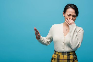 Disgusted asian woman pinching nose and showing no sign isolated on blue 