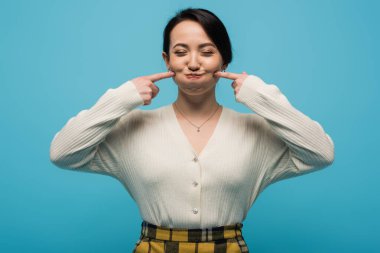 Young asian woman grimacing and touching cheeks isolated on blue 