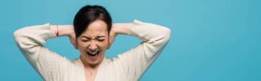 Aggressive asian woman screaming and covering ears isolated on blue, banner 