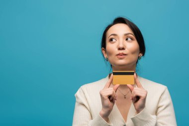 Thoughtful asian woman in cardigan holding credit card isolated on blue 