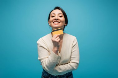 Positive asian woman in cardigan holding credit card isolated on blue 