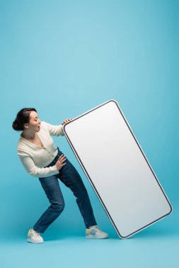 Shocked asian woman looking at big cellphone model on blue background