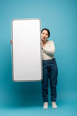 Shocked asian woman holding big smartphone model on blue background
