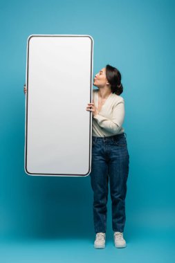 Side view of young asian woman pouting lips and holding big smartphone model on blue background