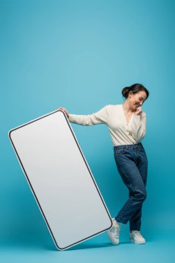 Cheerful asian woman standing near big smartphone mockup on blue background