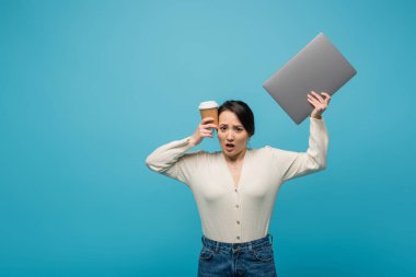 Worried asian model in cardigan holding laptop and coffee isolated on blue  clipart